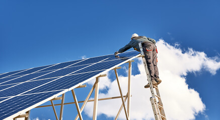 Wall Mural - Horizontal snapshot of a male worker wearing a uniform, standing on a ladder on the top of a solar plant, mounting solar modules on a sunny day. Clear blue sky and one white cloud on background