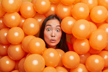 Photo of shocked brunette young Asian woman sticks head through orange inflated helium balloons has stunned expression celebrates holiday poses against decorated background. Festive event concept