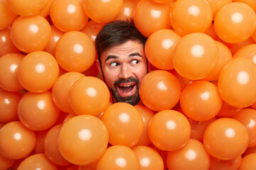 Wall Mural - Happy preparation to party. Positive bearded man sticks out head through inflating air balloons has festive mood concentrated aside smiles happily celebrates birthday gets ready for special holiday