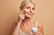 canvas print picture - Close up shot of middle aged beautiful woman applies anti aging cream on face undergoes beauty treatments cares about skin poses against beige background. Wrinkled female model with cosmetic product