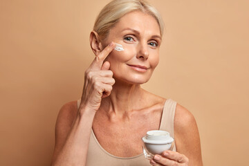 close up shot of middle aged beautiful woman applies anti aging cream on face undergoes beauty treat