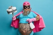 © WHstudio Leushin N - Confident bearded man housekeeper busy doing domestic work at home holds bottle of detergent and plunger involved in cleaning process wears superhero costume poses indoor with big tattooed belly