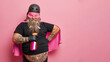 Bearded obese supernatural man with cleanser in hand wears superhero costume cleans everything at home poses against rosy background blank space for your advertising content. Housework concept