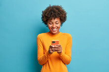 Online Lifestyle Concept. Cheerful Good Looking Woman With Afro Hair Sends Text Messages Via Mobile Phone Dressed Casually Searches Gifts For Holiday In Internet Uses Smartphone App Browses Webpage