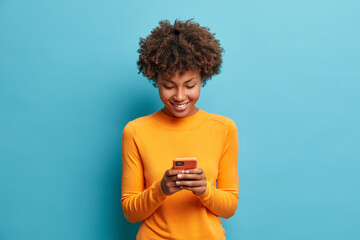 Wall Mural - Happy cheerful Afro American woman looks at screen of smart phone enjoys online chatting types text message surfs social networks dressed casually poses against blue background. Technology concept