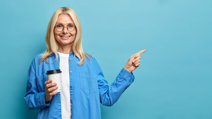 Smiling good looking senior woman feels happy and points to side shows nice advertisement holds takeaway coffee dressed in fashionable shirt isolated on blue background. People lifestyle concept