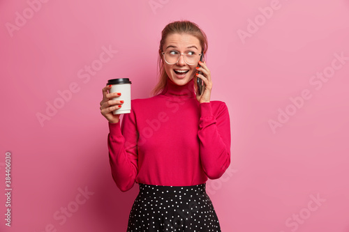 Shot of happy Caucasian woman with pony tail has interesting talk via mobile phone calls best friend holds take away coffee dressed in casual clothes isolated over rosy wall uses wireless connection