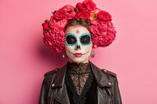 Indoor Shot Of Calm Woman With Closed Eyes Bright Makeup Prepares For Mexican Holiday Has Sugar Skull Wears Flower Wreath And Leather Jacket Celebrates Holiday To Honor Dead People Isolated On Pink