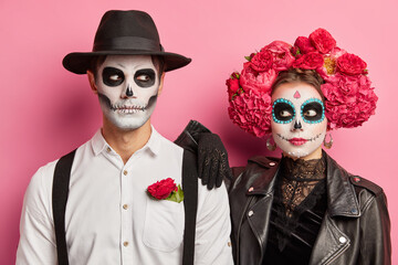 Horizontal shot of spooky couple dressed in day of dead costumes professional horrible makeup pose against pink background during halloween photoshoot. Celebration of traditional Mexican holiday