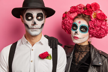 Close up shot of loving couple with spooky makeup pose during day of dead enjoy halloween party stand against rosy background wear mexican attire. Dead woman and man celebrate holiday in Mexico