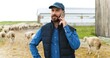 © VAKSMANV - Handsome smiled Caucasian male shepherd talking on mobile phone while flock of sheep walking on background. Man farmer speaking on telephone outdoors at shed.