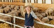 © VAKSMANV - Portrait of Caucasian blonde pretty girl with fair hair standing at fence in stable and smiling to camera. Sheep flock on background. Cute little kid farmer in hat laughing in livestock barn.