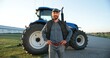 © VAKSMANV - Portrait of young Caucasian handsome man farmer in cap standing on the road in field and smiling to camera. Big tractor on background. Male worker in agricultural farm. Agriculture farming.