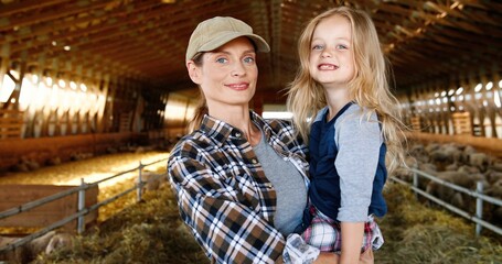 Wall Mural - Caucasian beautiful woman holding small cute pretty daughter on hands and smiling to camera in stable with sheep flock. Indoor. Little girl with lovely mother. Farming at countryside. Motherhood love.