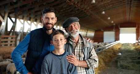 Wall Mural - Three male generations in green field at cattle farm. Caucasian grandfather, father and son smiling to camera and standing at stable with sheep flock. Shepherds. Old man farmer with son and grandson.