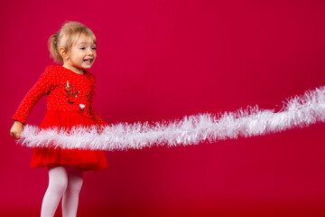 Pretty caucasian laughing little girl in red Xmas dress white stockings playing with an white christmas tinsel garland against red background. Holiday concept