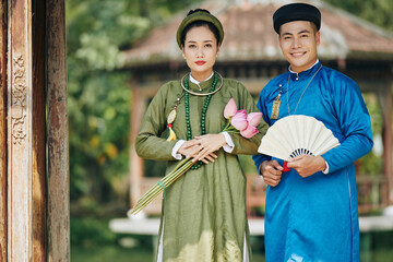 Sticker - Young Vietnamese couple in national costumes standing with lotus flowers and carved fan standing outdoors and smiling at camera