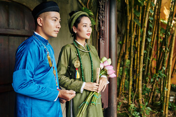 Sticker - Serious young Vietnamese couple in blue and green ao dai dresses standing at entrance doors of old temple