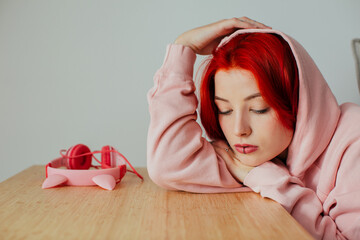 Wall Mural - Portrait of a serious teen girl with bright red hair, lip piercing and hoodie  resting head on desk looking down thinking