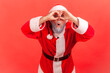 Portrait of curious funny gray bearded man in santa claus costume holding hands near eyes looking like through binocular and prying about holidays wishes. Indoor studio shot isolated on red background