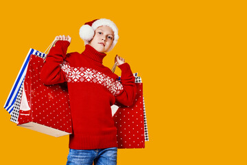 Cute boy in red sweater holding shopping bags with Christmas gifts and looking at camera on yellow background