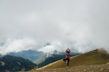 Wall Mural - Asana high up in cloudy mountains. Young woman doing yoga stance. Long shot