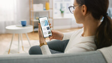 Young Woman At Home Uses Smartphone For Scrolling And Reading News About Technological Breakthroughs. She's Sitting On A Couch In Her Cozy Living Room. Over The Shoulder Shot