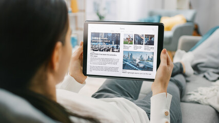 Wall Mural - Over the Shoulder Shot of a Young Woman at Home Lying on a Couch and Using Tablet Computer in Horizontal Landscape Mode for Reading News about Technology. Girl Using Touchscreen Device.