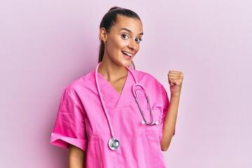 Wall Mural - Young hispanic woman wearing doctor uniform and stethoscope smiling with happy face looking and pointing to the side with thumb up.