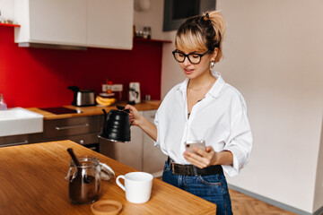 Joyful young woman preparing coffee. Indoor shot of girl in glasses holding black kettle in kitchen.