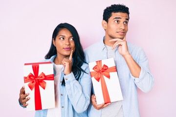 Wall Mural - Beautiful latin young couple holding gift for anniversary celebration serious face thinking about question with hand on chin, thoughtful about confusing idea