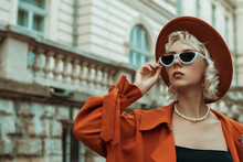 Autumn Outdoor Portrait Of Fashionable Blonde Woman Wearing Black, White Cat Eye Sunglasses, Pearl Necklace, Orange Hat, Trench Coat, Posing In Street Of European City. Copy, Empty Space For Text