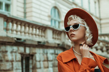 Fashionable Blonde Woman Wearing Black, White Cat Eye Sunglasses, Pearl Necklace, Ring, Orange Hat, Trench Coat, Posing In Street Of European City. Copy, Empty Space For Text