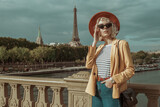Fototapeta Paryż - Outdoor portrait of stylish woman wearing autumn outfit: orange hat, black cat eye sunglasses, yellow blazer, striped t-shirt, red belt, wrist watch, posing in street of Paris