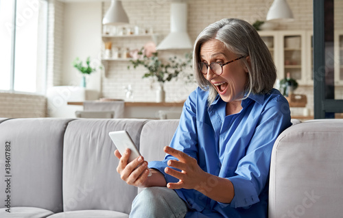 Fotografía Amazed happy mature older 60s woman, excited customer holding smartphone using mobile app feeling great positive surprise reaction receiving gift reading sms on cell phone sitting on couch at home