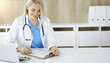 © Iryna - Woman-doctor at work while sitting at the desk in sunny clinic. Blonde cheerful physician filling up medication history record form