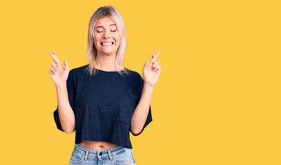 Young beautiful blonde woman wearing casual t-shirt gesturing finger crossed smiling with hope and eyes closed. luck and superstitious concept.