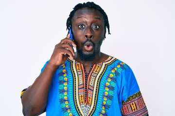 Poster - Young african american man with braids wearing ethnic tshirt having conversation talking on the smartphone scared and amazed with open mouth for surprise, disbelief face