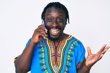 Canvas Print - Young african american man with braids wearing ethnic tshirt having conversation talking on the smartphone celebrating victory with happy smile and winner expression with raised hands