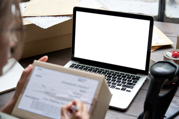 Female seller, internet shop small business owner holding ecommerce shipping box checking online store dropshipping order on mock up white blank laptop screen. E commerce post shipping delivery.