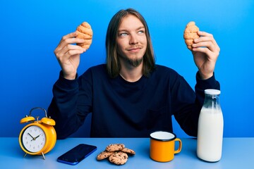 Wall Mural - Handsome caucasian man with long hair eating croissant for breakfast smiling looking to the side and staring away thinking.