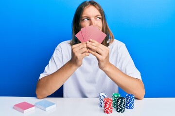 Wall Mural - Handsome caucasian man with long hair playing gambling poker covering face with cards smiling looking to the side and staring away thinking.