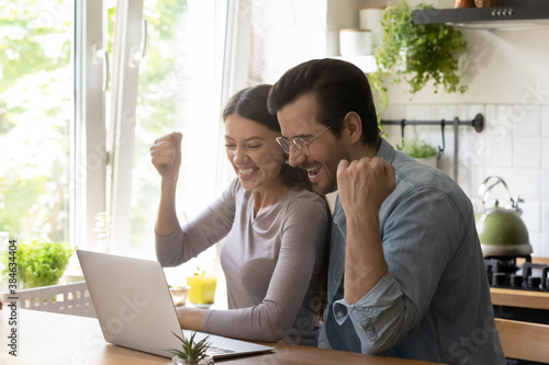Fotografía Overjoyed young married couple making yes gesture, celebrating online lottery win or family success achievement, looking at computer screen, relaxing at modern kitchen table, good luck concept