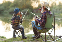 Careful Grandpa With Grey Beard In Straw Hat Sitting On Folding Chairs With His Grandchild On Fishing And Drinking Tea.