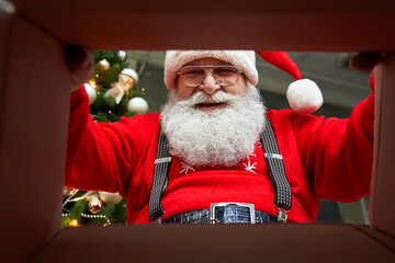 Wall Mural - Happy old bearded Santa Claus wearing hat packing present looking inside cardboard box wrapping gift preparing package delivery on xmas eve. Merry Christmas surprise concept, close up view from below.