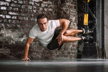 Wall Mural - Serious sportsman with legs on TRX ropes looking at camera during intense suspension workout against rough brick wall