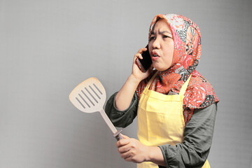 An Asian Muslim woman looks angry when she calls while holding a spatula to cook. for bussines from home