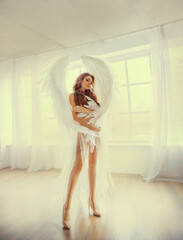 Wall Mural - Young beautiful woman angel with huge white wings stands in the room near the window.