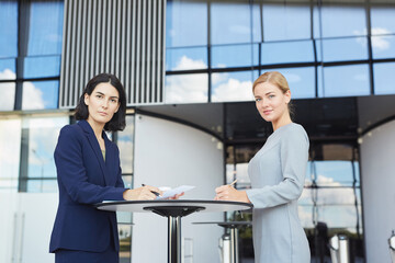 Wall Mural - Side view portrait of two businesswomen smiling at camera while standing by cafe table in airport or office building, copy space