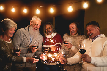 Wall Mural - Happy multiracial people celebrating new year's eve drinking wine at home - Mature friends having fun with fireworks sparklers outdoor - Holiday and party concept - Soft focus on african woman